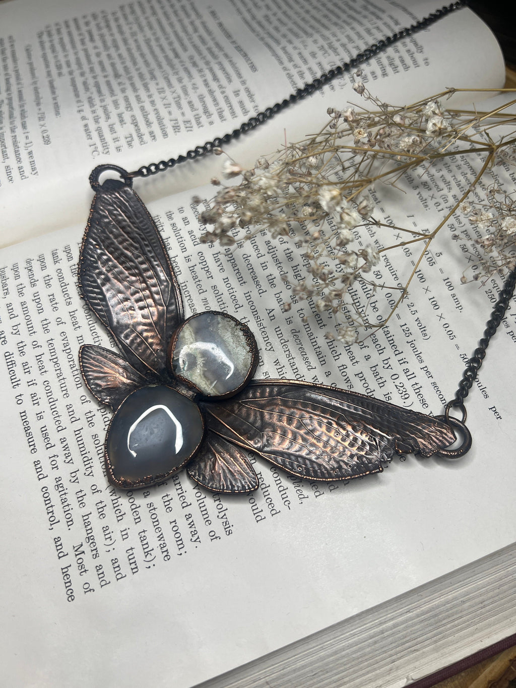 May include: A copper and black necklace with a dragonfly design. The dragonfly has two sets of wings, each with a different texture. The wings are accented with two round, clear stones with black and white patterns. The necklace is resting on a book with text.