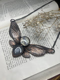 May include: A copper and black necklace with a dragonfly design. The dragonfly has two sets of wings, each with a different texture. The wings are accented with two round, clear stones with black and white patterns. The necklace is resting on a book with text.