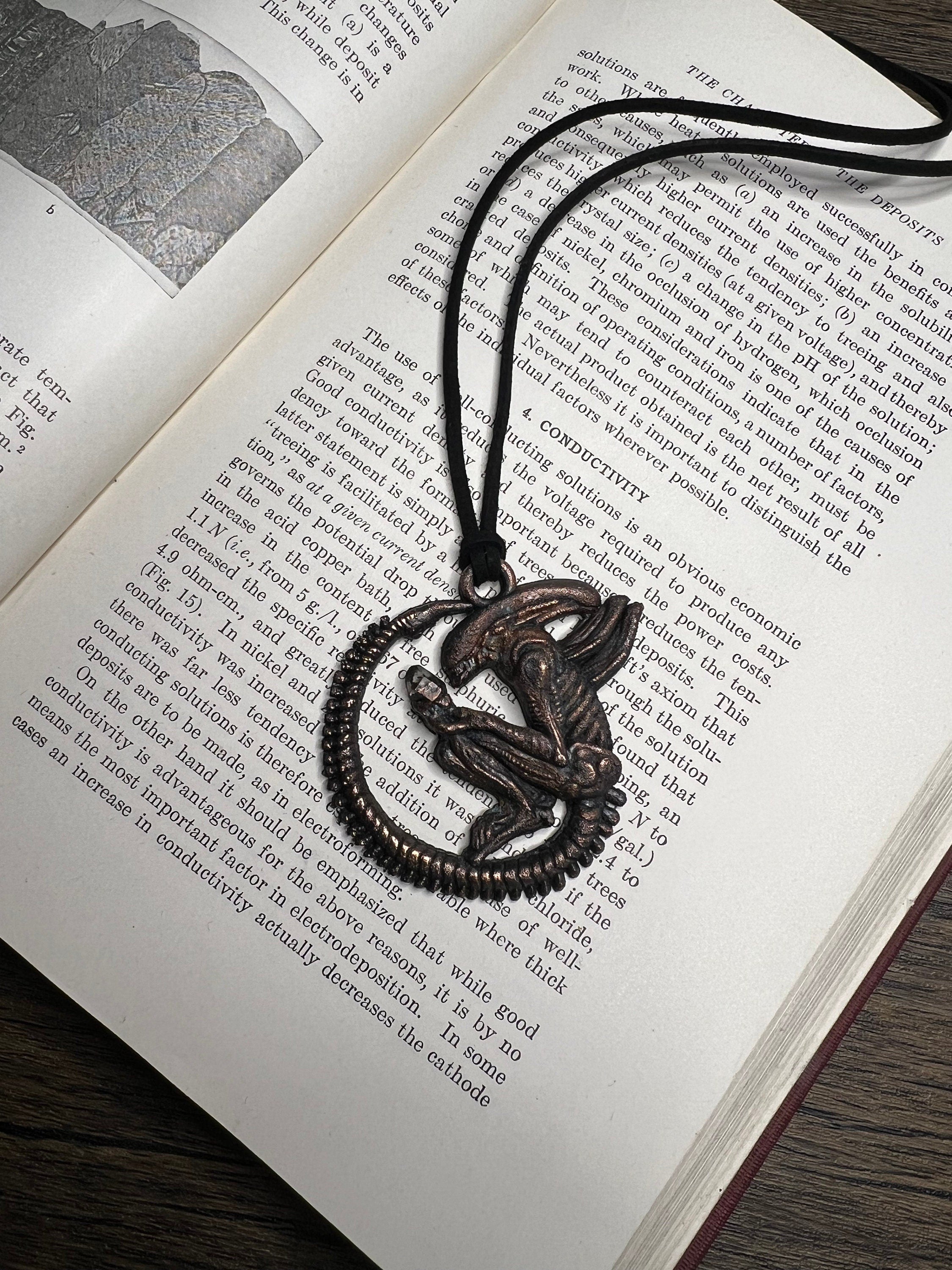May include: A bronze-colored pendant featuring a detailed, skeletal creature with a long tail and wings. The pendant is hanging from a black cord and is surrounded by a circular, metal frame.