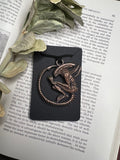 May include: A black and bronze pendant featuring a detailed alien creature with a diamond-shaped gem on its chest. The pendant is hanging from a black cord and is displayed on a black background.