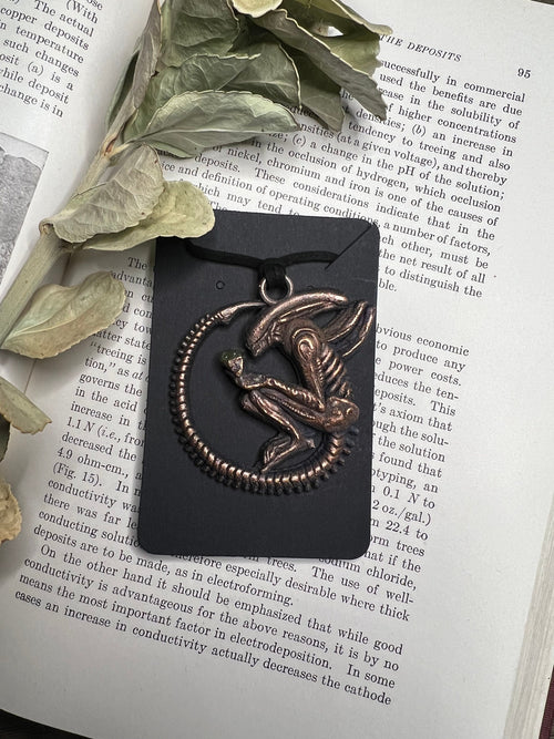 May include: A black and bronze pendant featuring a detailed alien creature with a diamond-shaped gem on its chest. The pendant is hanging from a black cord and is displayed on a black background.