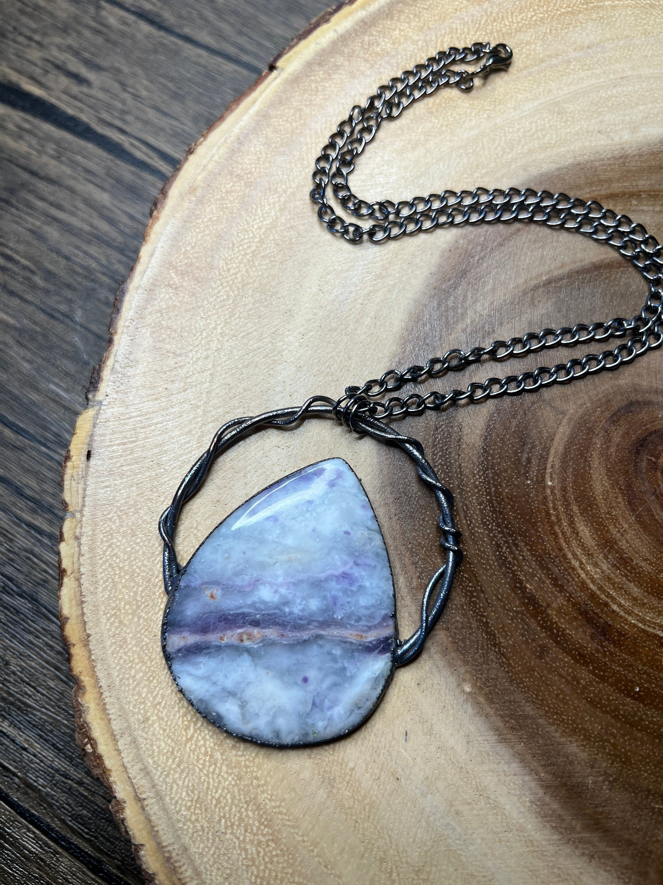 May include: A silver chain necklace with a teardrop-shaped purple and white stone pendant. The stone is set in a silver wire frame.
