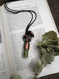 May include: A close-up of a copper and silver necklace with a mushroom pendant. The pendant is filled with green crystals and is hanging from a black cord. The necklace is resting on a page of a book with text about the character of deposits.