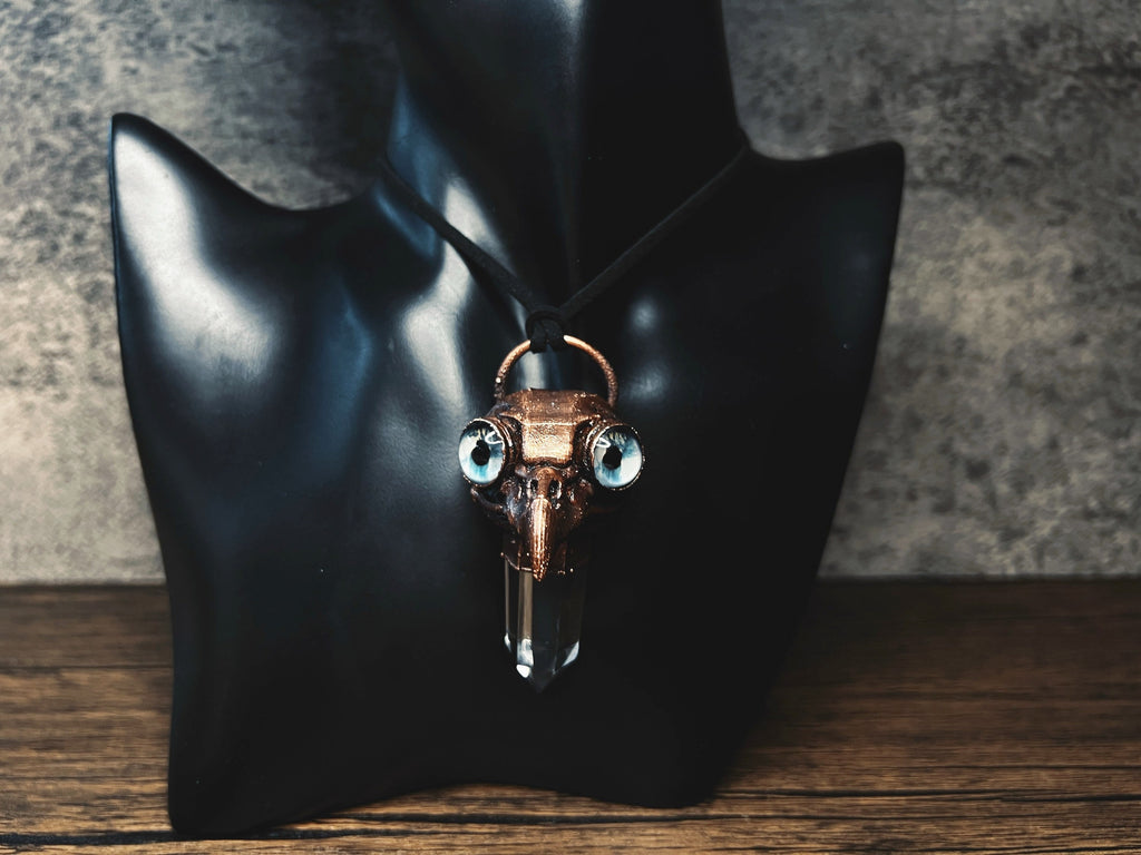 May include: A copper-colored pendant necklace with a crystal point and two blue eyes. The pendant is shaped like a bird skull.