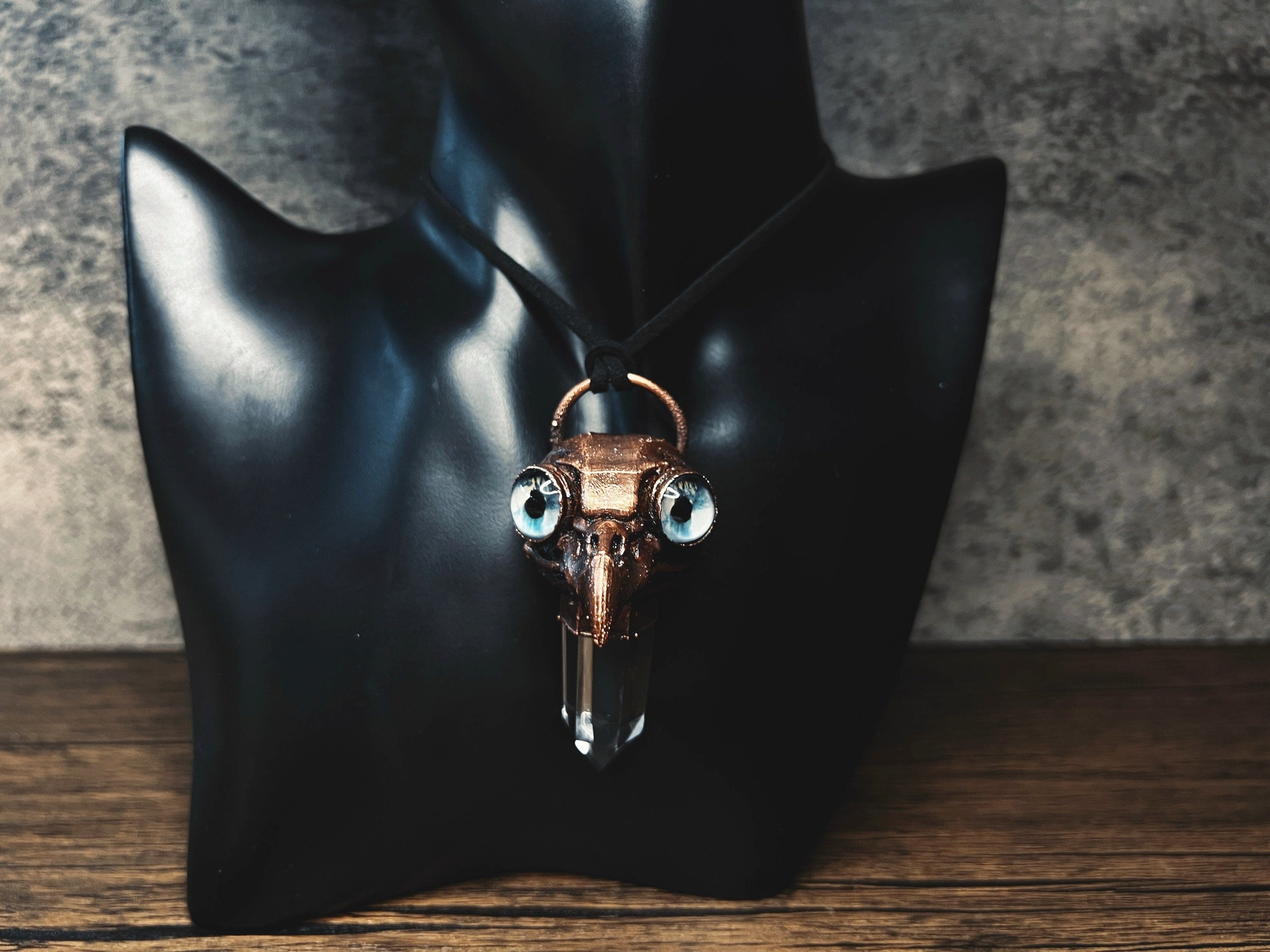 May include: A copper-colored pendant necklace with a crystal point and two blue eyes. The pendant is shaped like a bird skull.