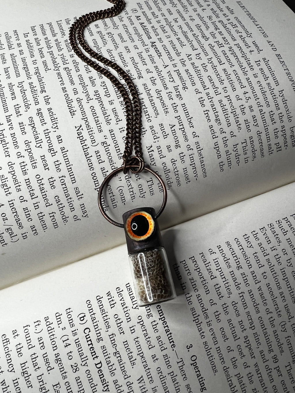 May include: A copper chain necklace with a small glass vial pendant. The vial is filled with a light brown substance and has a black and orange top. The necklace is lying on a page of a book about electroplating and electroforming.