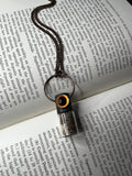 May include: A copper chain necklace with a small glass vial pendant. The vial is filled with a light brown substance and has a black and orange top. The necklace is lying on a page of a book about electroplating and electroforming.