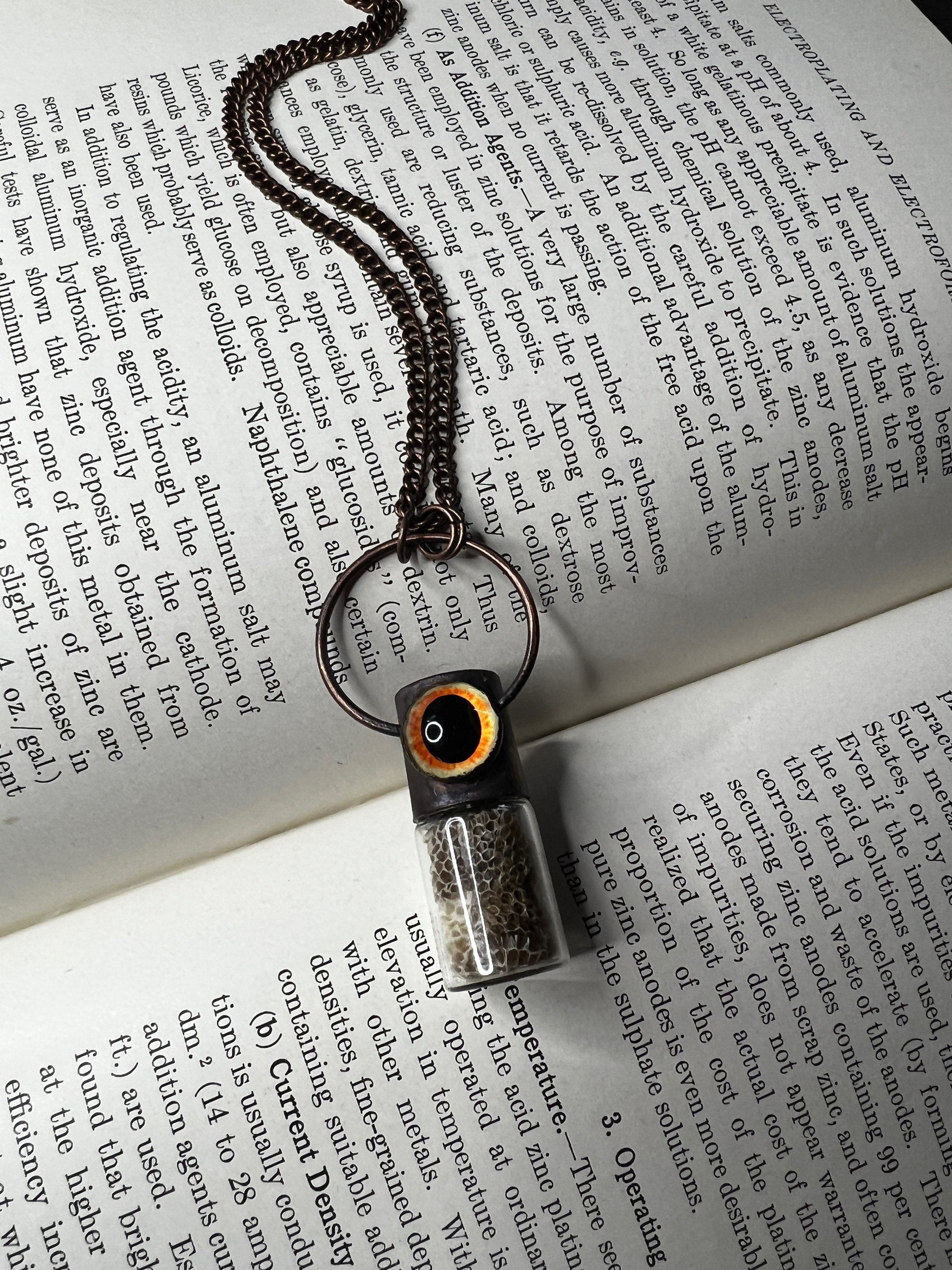 May include: A copper chain necklace with a small glass vial pendant. The vial is filled with a light brown substance and has a black and orange top. The necklace is lying on a page of a book about electroplating and electroforming.