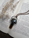 May include: A black and silver necklace with a pendant featuring a glass bottle with a brown liquid inside and a black and silver creature with a blue eye. The necklace is on top of a book with text about electrochemistry.