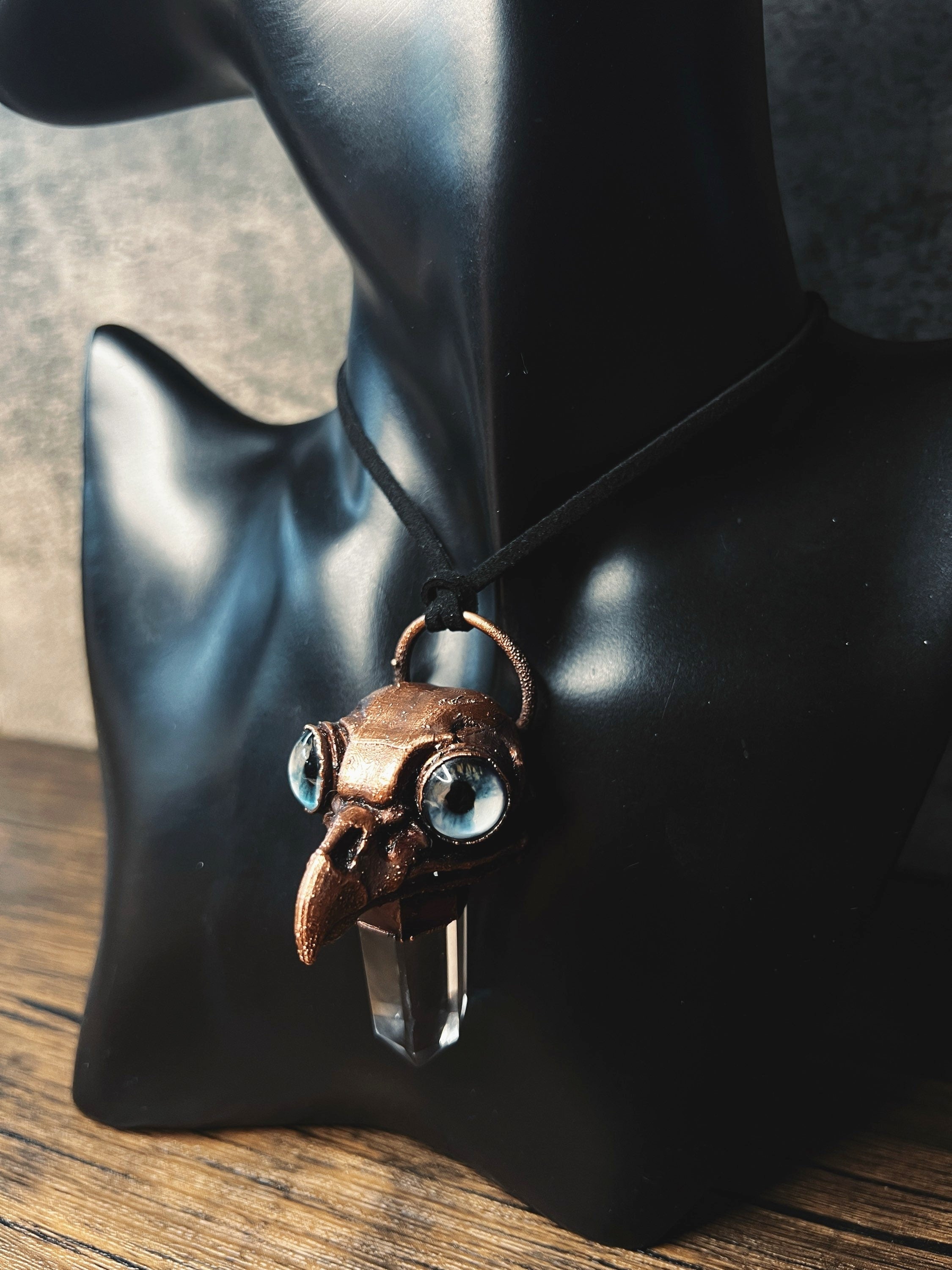 May include: A copper-colored pendant necklace with a bird skull design and blue glass eyes. The pendant is attached to a clear quartz crystal and hangs from a black cord.