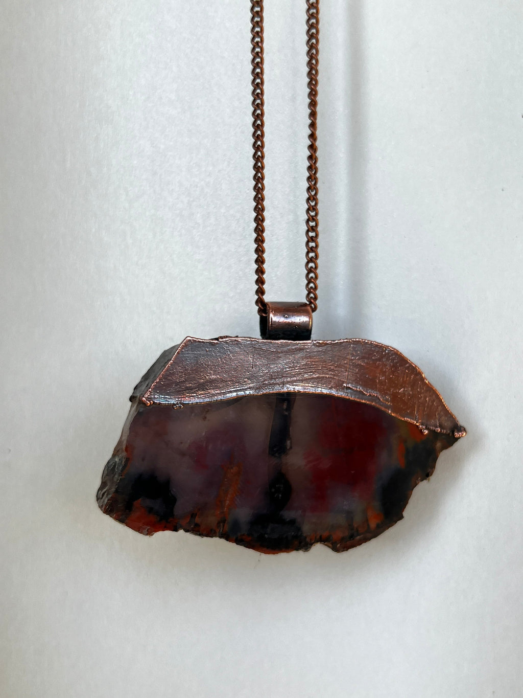 May include: A copper chain necklace with a pendant made from a slice of agate. The agate is a mix of red, black, and white colors. The copper chain is a simple, delicate design.