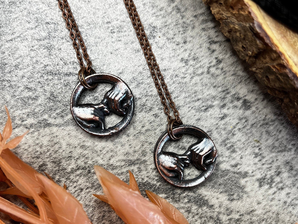 May include: Two copper necklaces with a circular pendant featuring a pair of hands clasped together. The pendants are a dark silver color.