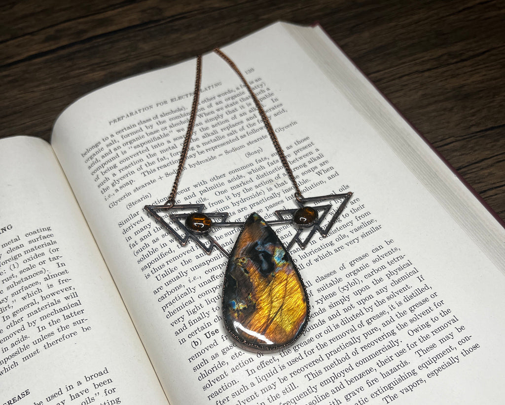 May include: A necklace with a teardrop-shaped pendant featuring a colorful butterfly wing encased in resin. The pendant is suspended from a metal chain with two triangular charms, each adorned with a brown gemstone.