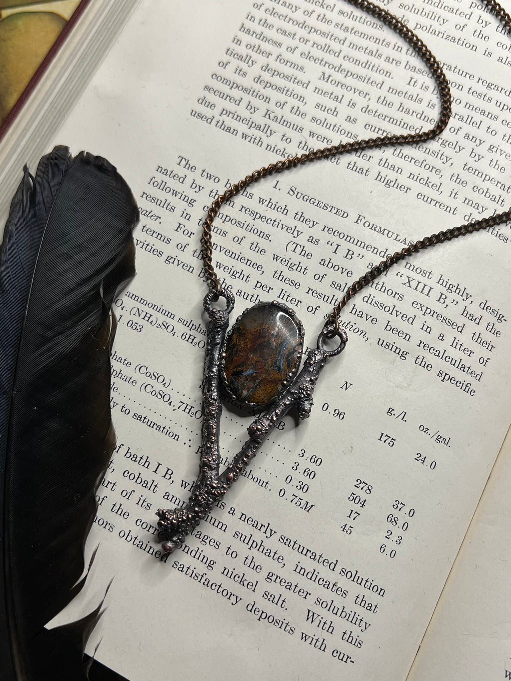 May include: A silver metal tree branch with a brown and orange agate stone pendant. The branch is decorated with small, textured details. The text "XIII B" is visible on the page behind the branch.