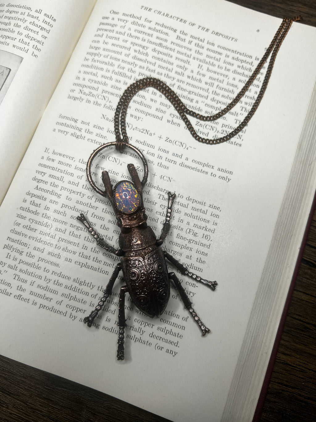 May include: A bronze beetle charm with a dark blue iridescent stone on its back, hanging from a double chain necklace. The charm is detailed with intricate designs and is resting on a page of a book with text about metal deposits.