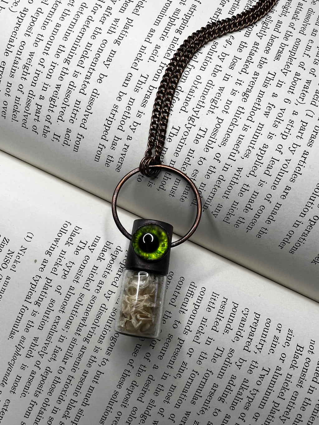 May include: A copper chain necklace with a glass vial pendant. The vial contains small white pieces of bone and has a black cap with a green eye on top. The eye has a black pupil and a green iris.