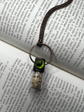 May include: A copper chain necklace with a glass vial pendant. The vial contains small white pieces of bone and has a black cap with a green eye on top. The eye has a black pupil and a green iris.