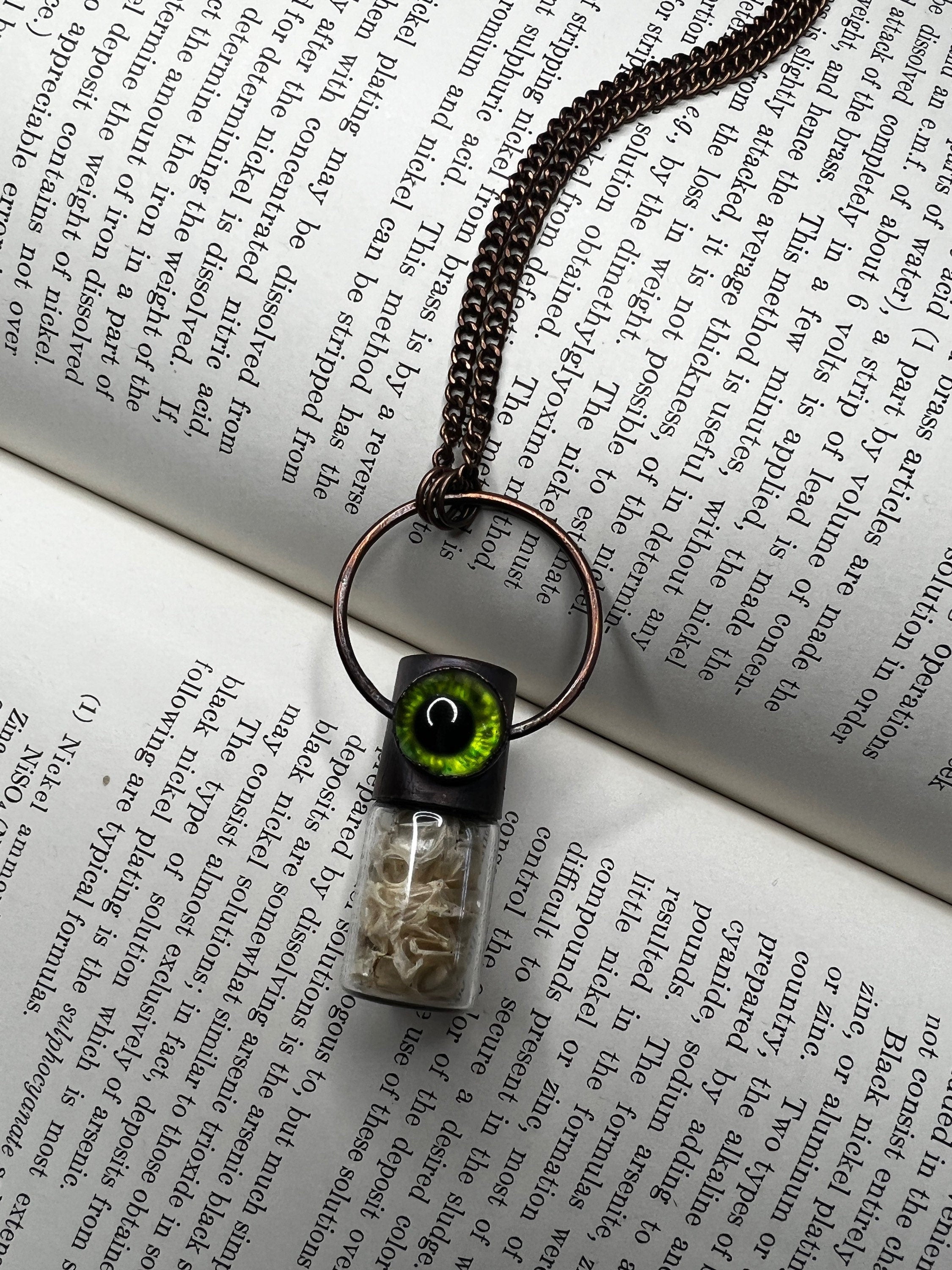May include: A copper chain necklace with a glass vial pendant. The vial contains small white pieces of bone and has a black cap with a green eye on top. The eye has a black pupil and a green iris.
