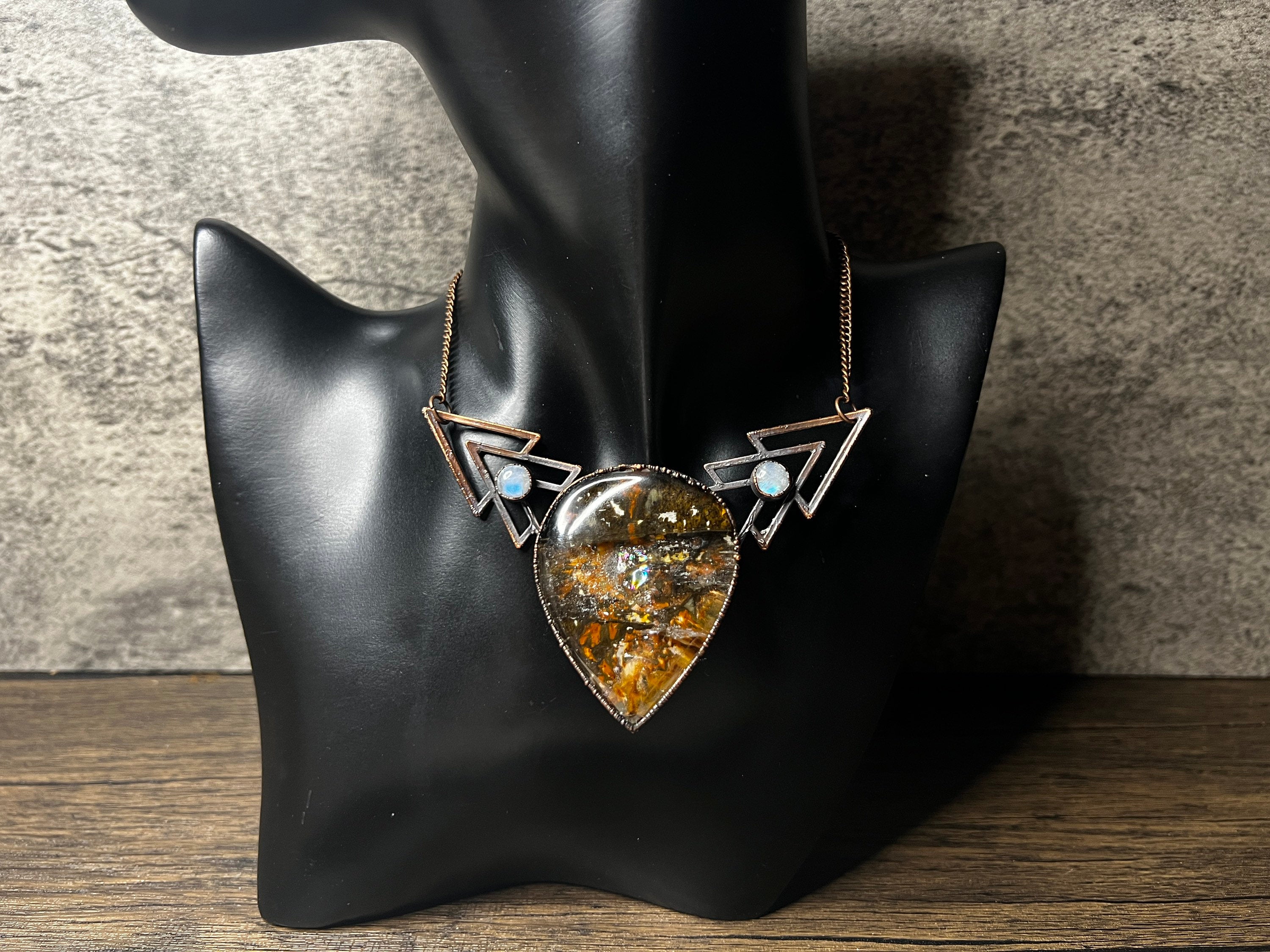 May include: A unique necklace featuring a large, teardrop-shaped pendant with a textured, amber-colored center. The pendant is framed in a silver metal and flanked by geometric silver accents, each with a small, round, white stone. The necklace is displayed on a black mannequin.