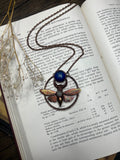 May include: A copper and blue butterfly pendant necklace with a blue gemstone. The pendant is surrounded by a copper circle with the text "Cobalt Sulphate" written on it. The necklace is on a book with text about cobalt and nickel.