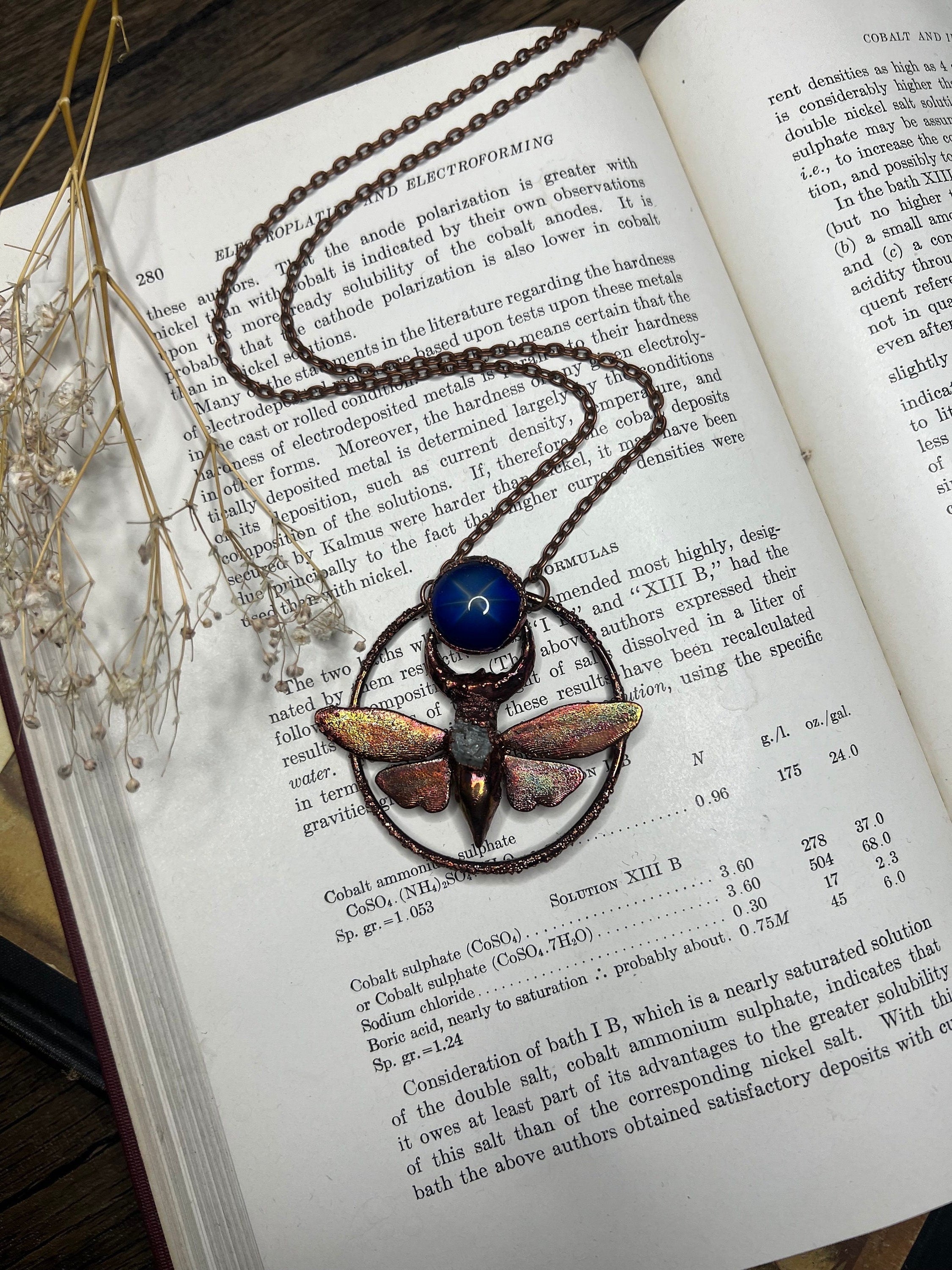 May include: A copper and blue butterfly pendant necklace with a blue gemstone. The pendant is surrounded by a copper circle with the text "Cobalt Sulphate" written on it. The necklace is on a book with text about cobalt and nickel.