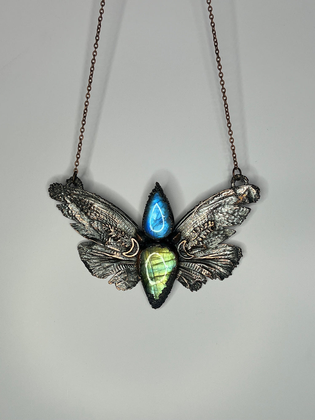 May include: A copper butterfly pendant necklace with a blue and green labradorite gemstone. The wings of the butterfly are detailed with intricate designs.