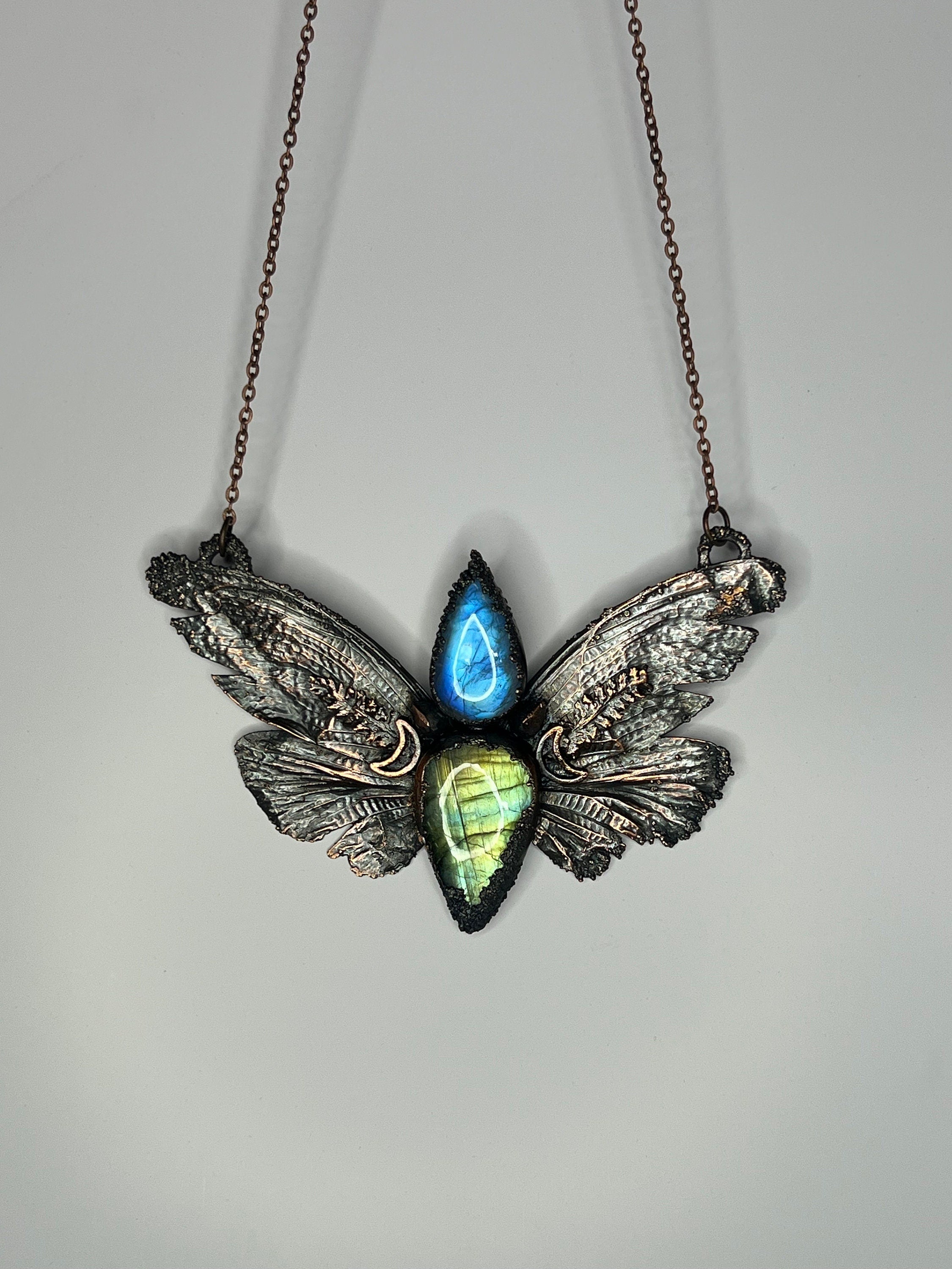 May include: A copper butterfly pendant necklace with a blue and green labradorite gemstone. The wings of the butterfly are detailed with intricate designs.
