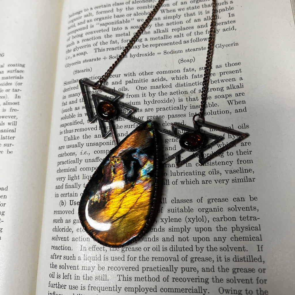 May include: A close-up of a necklace with a copper chain and three triangular pendants. The pendants are decorated with iridescent glass stones in shades of blue, green, yellow, and orange. The text "Soap" is visible on the page in the background.