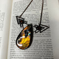May include: A close-up of a necklace with a copper chain and three triangular pendants. The pendants are decorated with iridescent glass stones in shades of blue, green, yellow, and orange. The text "Soap" is visible on the page in the background.
