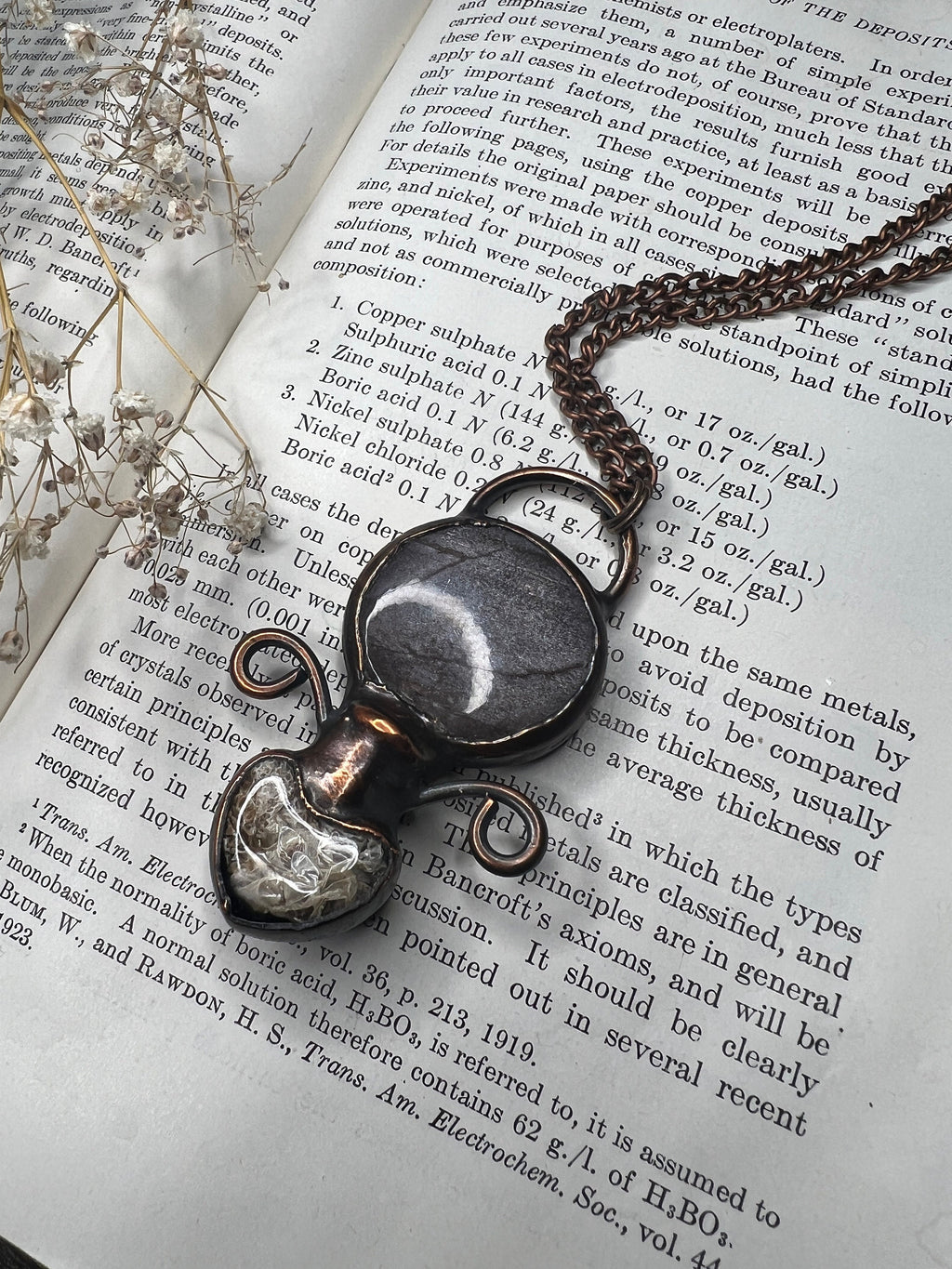 May include: A copper chain necklace with a pendant made of a round, gray and white stone encased in a clear glass dome. The pendant is attached to the chain with a copper wire loop. The necklace is lying on a page of a book with text about electrodeposition.
