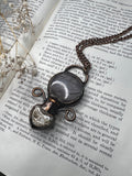 May include: A copper chain necklace with a pendant made of a round, gray and white stone encased in a clear glass dome. The pendant is attached to the chain with a copper wire loop. The necklace is lying on a page of a book with text about electrodeposition.