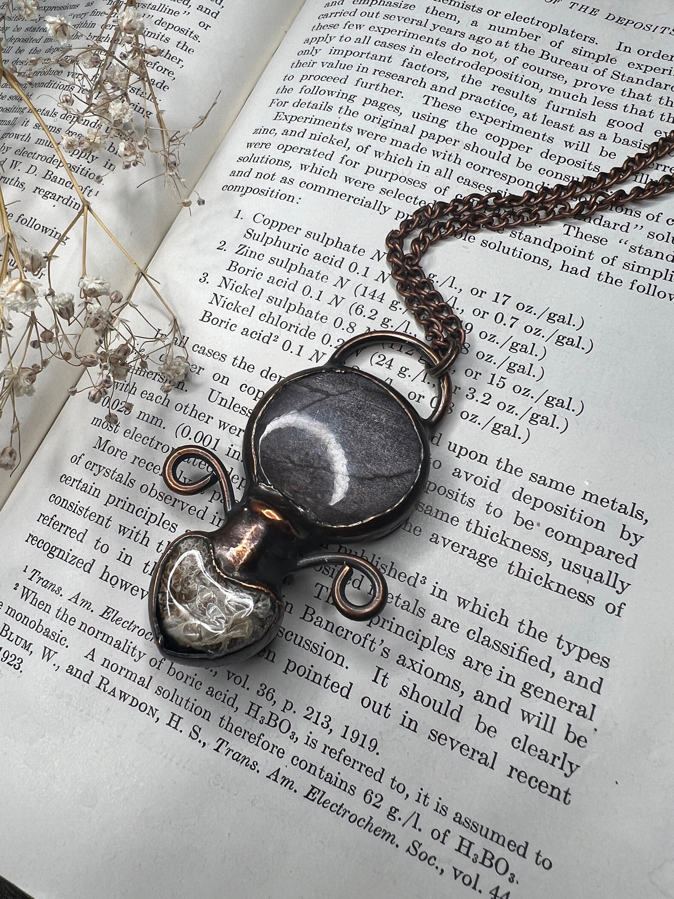 May include: A copper chain necklace with a pendant made of a round, gray and white stone encased in a clear glass dome. The pendant is attached to the chain with a copper wire loop. The necklace is lying on a page of a book with text about electrodeposition.