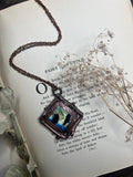 May include: A copper necklace with a square pendant featuring a blue and white iridescent stone. The pendant is set in a copper frame with a rough, textured finish. The necklace is lying on a book with text about fairies and children.