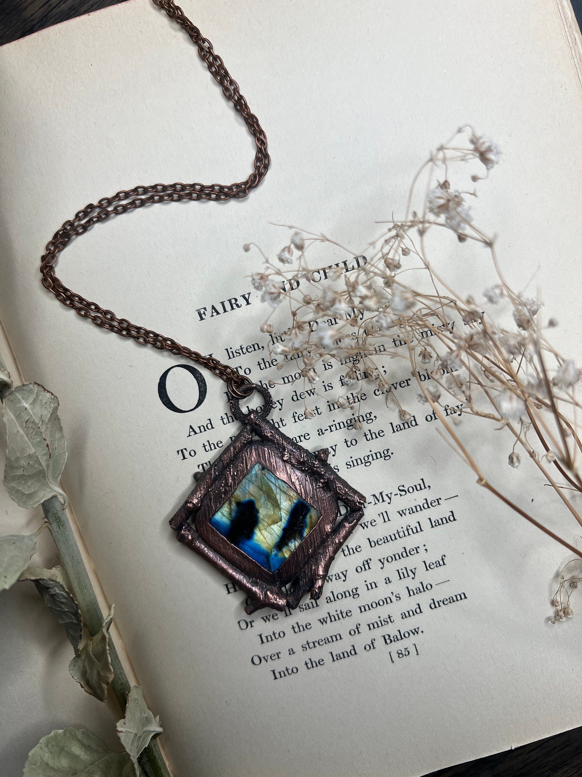 May include: A copper necklace with a square pendant featuring a blue and white iridescent stone. The pendant is set in a copper frame with a rough, textured finish. The necklace is lying on a book with text about fairies and children.
