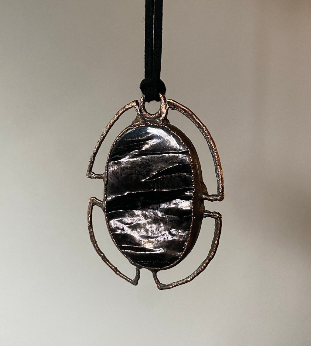 May include: A copper pendant with a black and silver oval stone set in a geometric frame. The pendant hangs from a black cord.