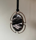 May include: A copper pendant with a black and silver oval stone set in a geometric frame. The pendant hangs from a black cord.