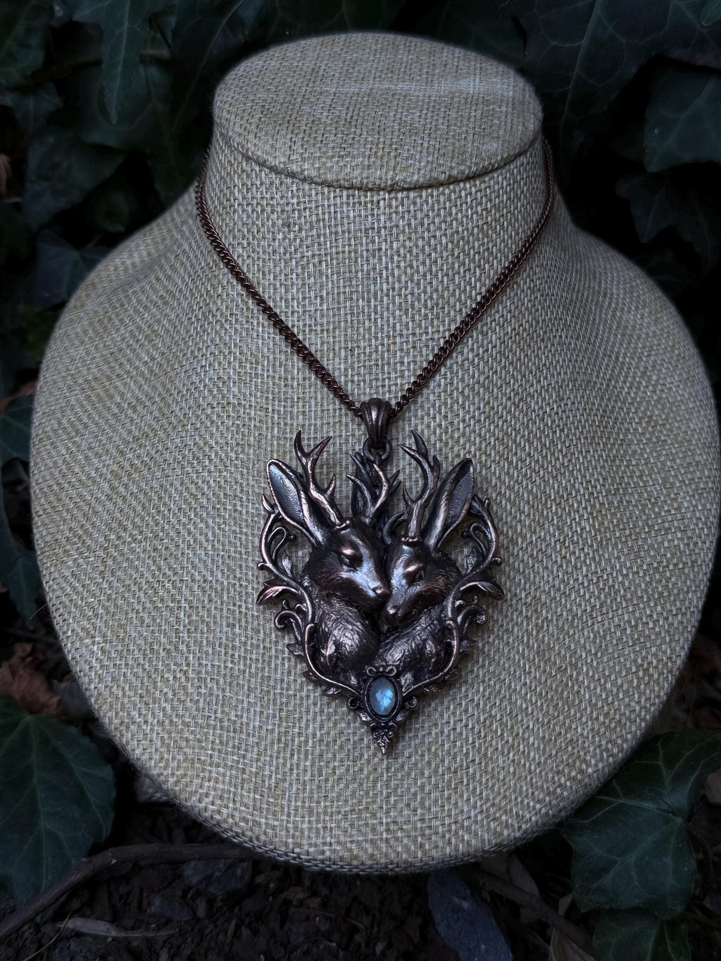 May include: A copper-toned pendant necklace featuring two jackalopes with prominent antlers, surrounded by intricate filigree leaf detailing and a small, iridescent blue cabochon stone at the base, suspended from a matching copper-colored chain.