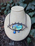 May include: A necklace featuring a large, oval-shaped labradorite stone with blue and gold iridescence, set in a dark, textured metal frame shaped like an eye with spikes. Two smaller, teardrop-shaped labradorite stones hang from the bottom.