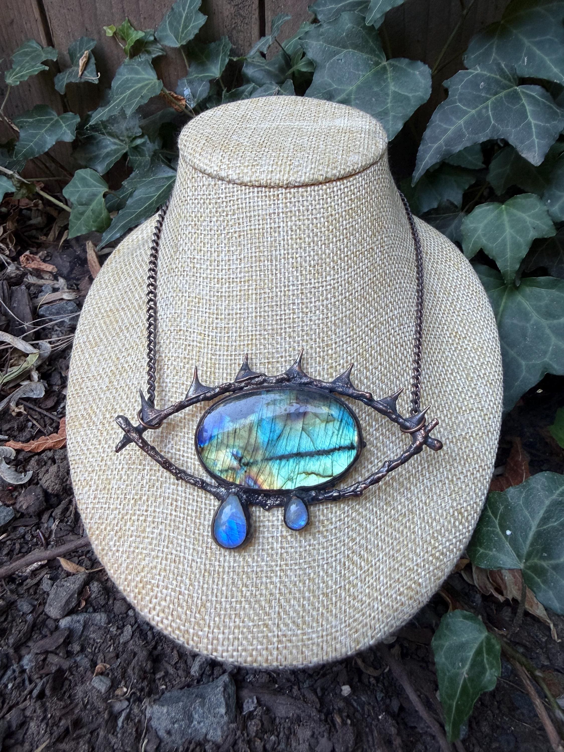 May include: A necklace featuring a large, oval-shaped labradorite stone with blue and gold iridescence, set in a dark, textured metal frame shaped like an eye with spikes. Two smaller, teardrop-shaped labradorite stones hang from the bottom.