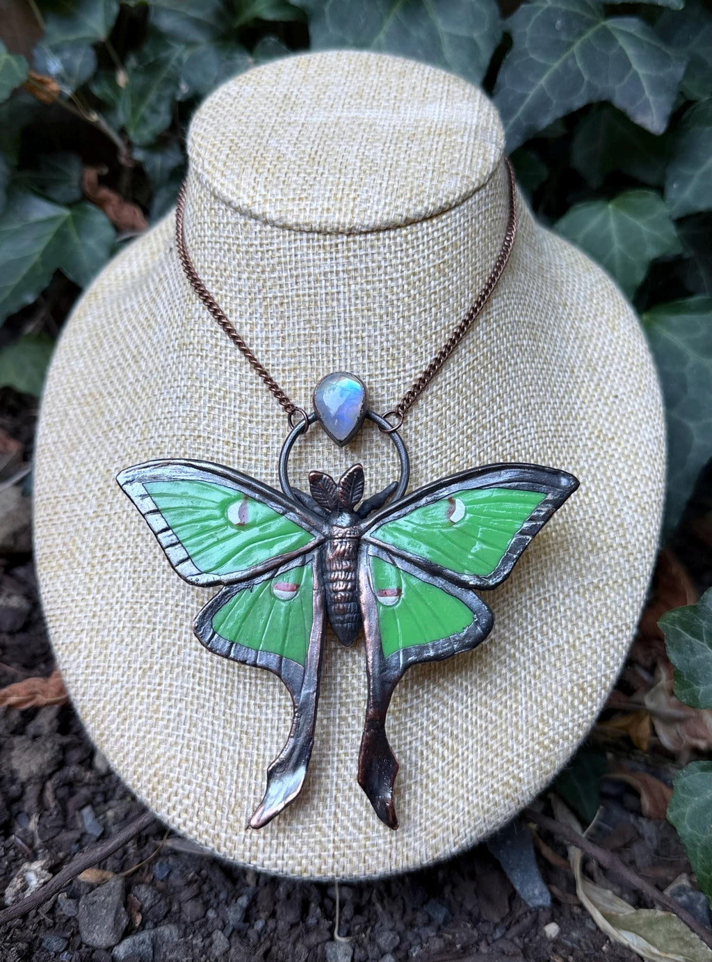 May include: A necklace featuring a large, green moth pendant with dark metallic edges and a small, iridescent teardrop-shaped stone set above the moth. The pendant hangs from a thin, dark copper-colored chain, showcasing detailed wing patterns and a textured body.