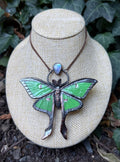 May include: A necklace featuring a large, green moth pendant with dark metallic edges and a small, iridescent teardrop-shaped stone set above the moth. The pendant hangs from a thin, dark copper-colored chain, showcasing detailed wing patterns and a textured body.