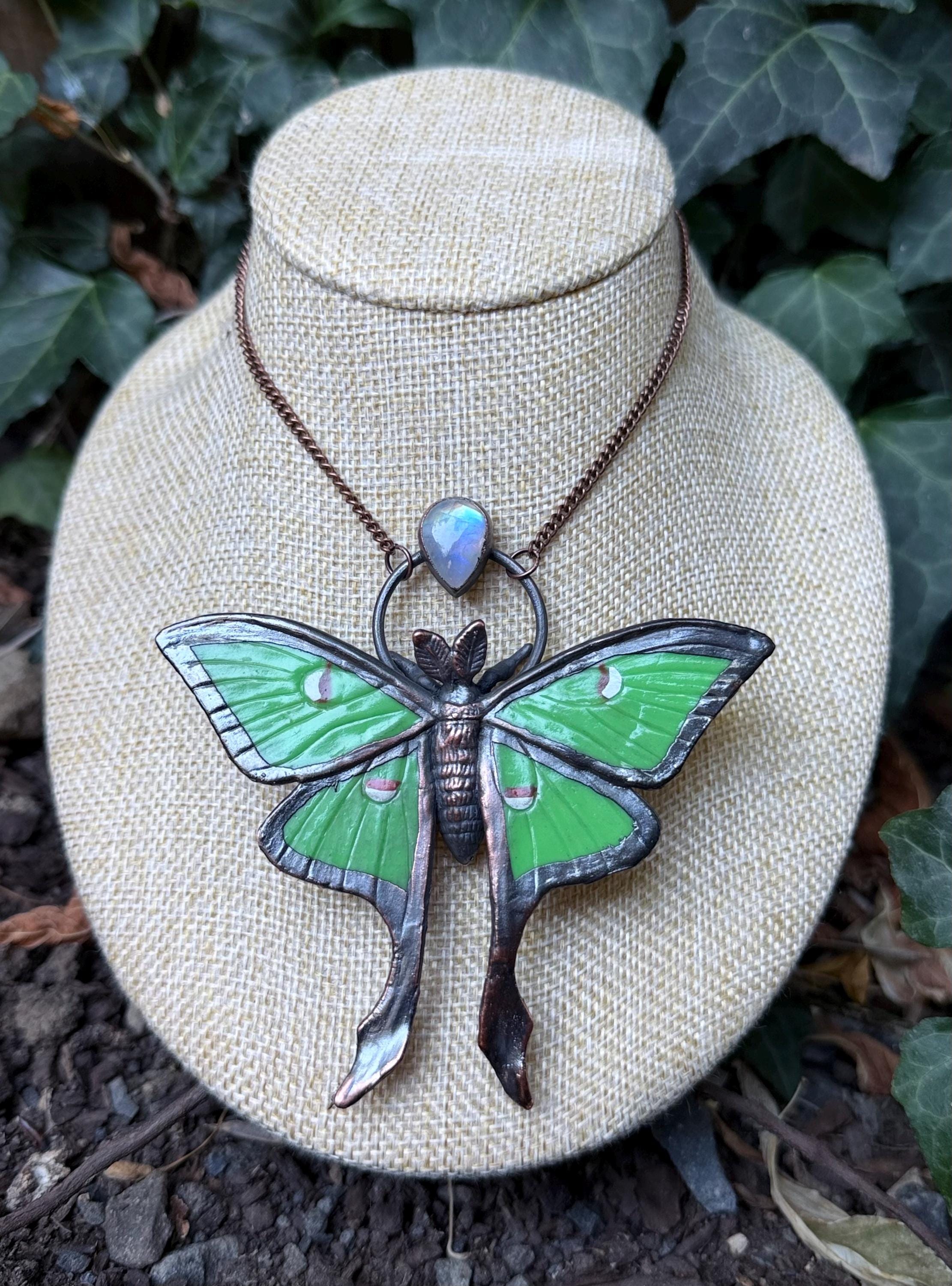 May include: A necklace featuring a large, green moth pendant with dark metallic edges and a small, iridescent teardrop-shaped stone set above the moth. The pendant hangs from a thin, dark copper-colored chain, showcasing detailed wing patterns and a textured body.