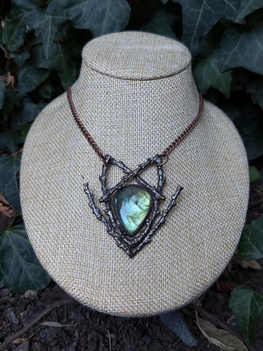 May include: A necklace featuring a teardrop-shaped labradorite gemstone with iridescent blue and green flashes, set within a dark, textured metal frame resembling intertwined twigs and branches, suspended from a copper-toned chain.