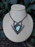 May include: A necklace featuring a teardrop-shaped labradorite gemstone with iridescent blue and green flashes, set within a dark, textured metal frame resembling intertwined twigs and branches, suspended from a copper-toned chain.