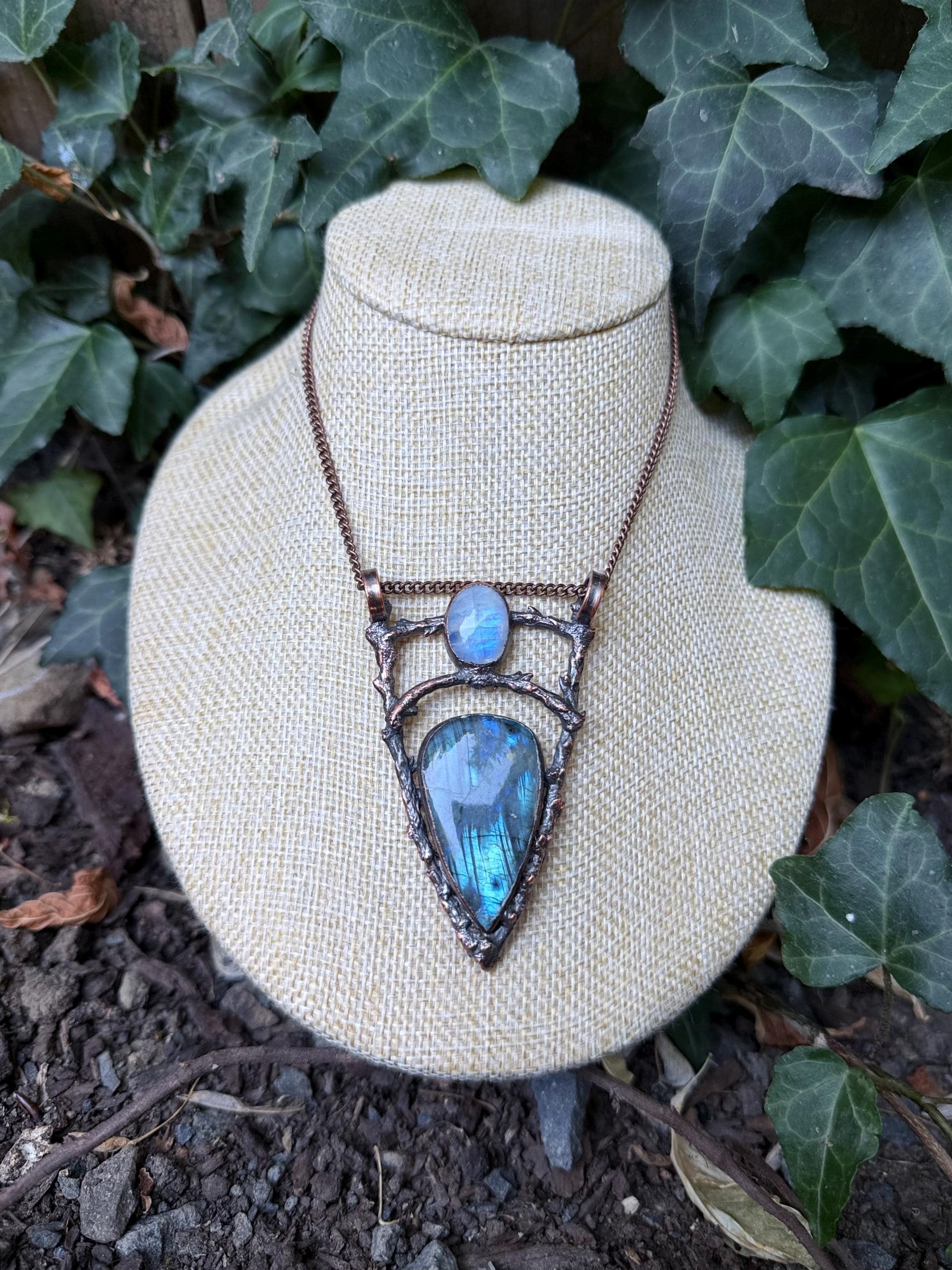 May include: A copper-toned necklace featuring a triangular pendant with a textured, branch-like frame. The pendant holds a large, pear-shaped labradorite stone with blue iridescence and a smaller, oval-shaped moonstone set above it.