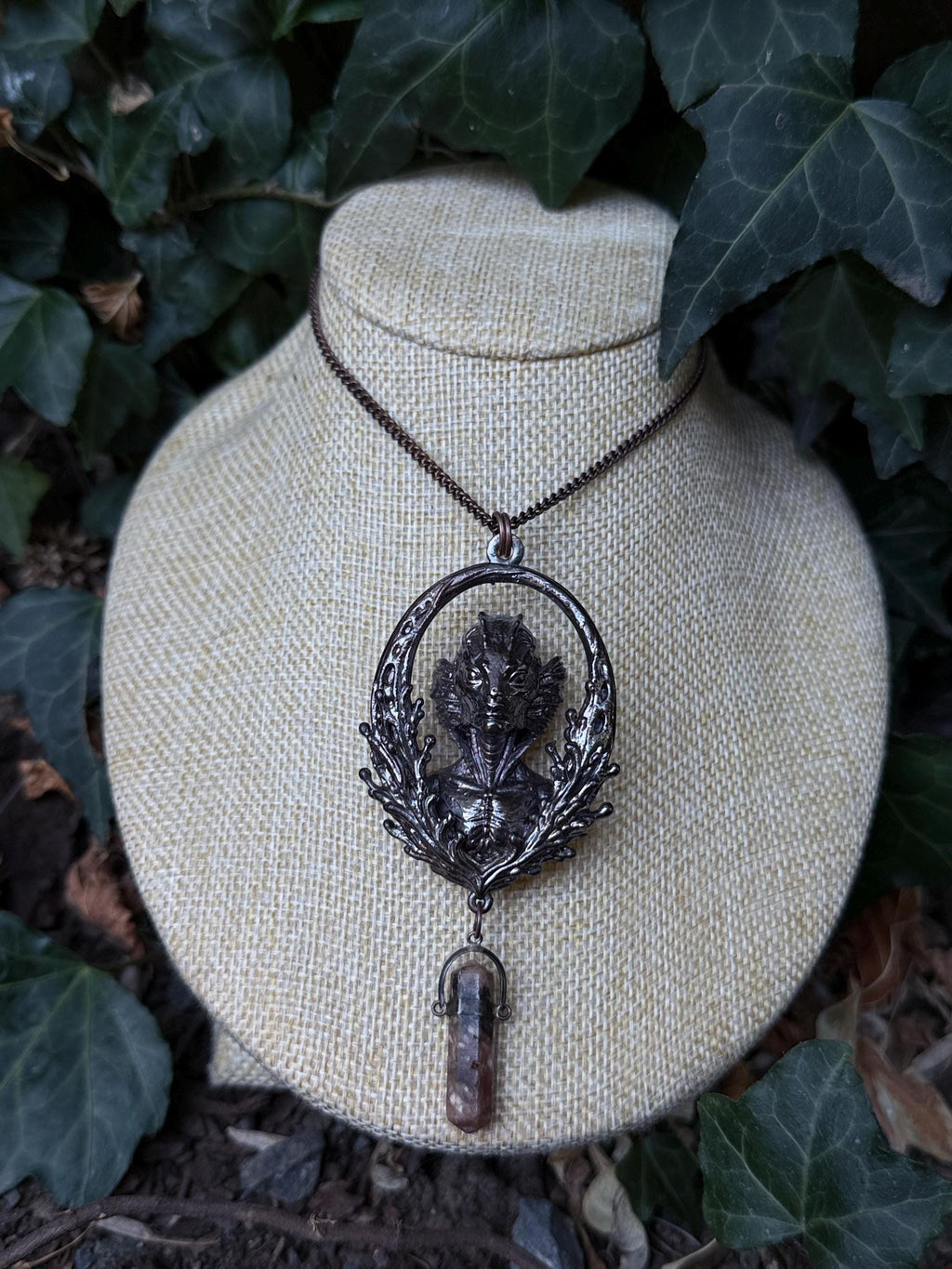 May include: A dark metallic pendant necklace featuring an oval frame with an intricate aquatic creature relief at the center. The pendant is suspended from a thin chain and includes a small, dangling, translucent brownish crystal point at the base.