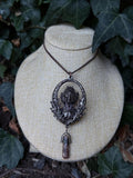 May include: A dark metallic pendant necklace featuring an oval frame with an intricate aquatic creature relief at the center. The pendant is suspended from a thin chain and includes a small, dangling, translucent brownish crystal point at the base.