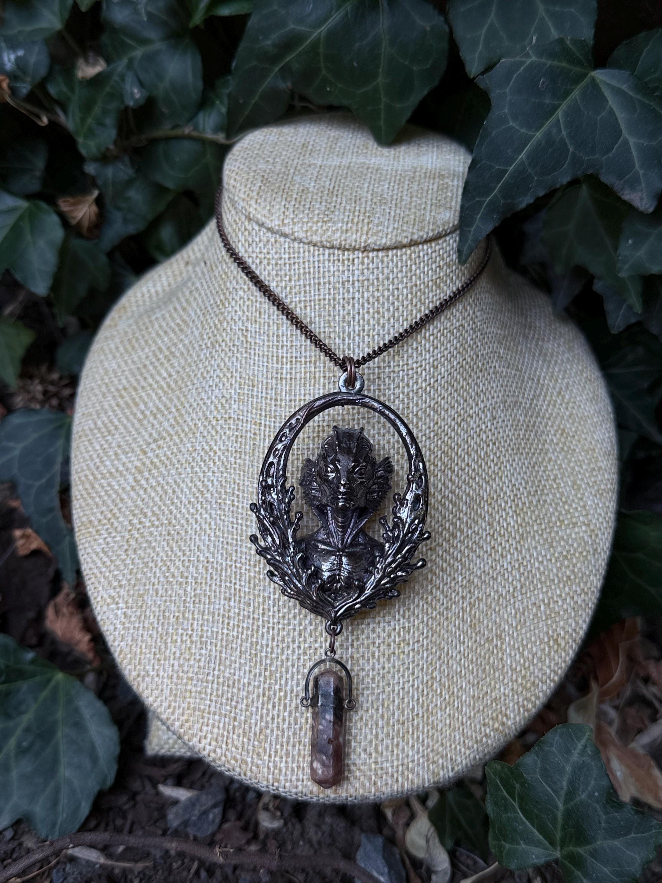 May include: A dark metallic pendant necklace featuring an oval frame with an intricate aquatic creature relief at the center. The pendant is suspended from a thin chain and includes a small, dangling, translucent brownish crystal point at the base.