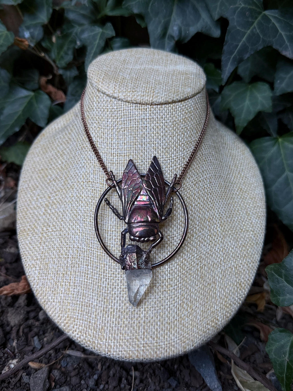 May include: A copper-toned necklace featuring a circular frame with a central insect pendant. The insect has iridescent wings and holds a clear, pointed quartz crystal point at the bottom. The piece hangs from a matching copper-colored metal chain.