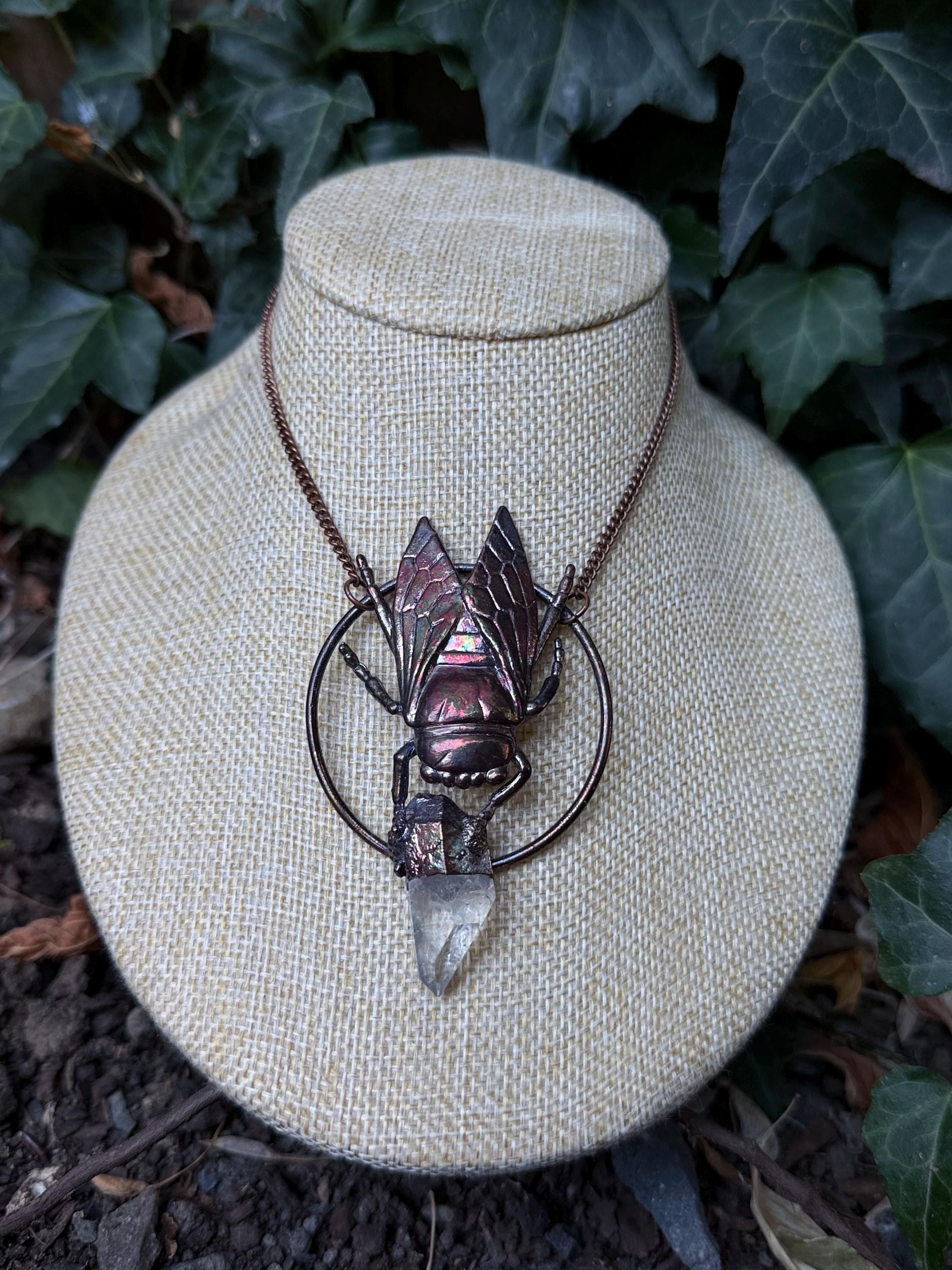 May include: A copper-toned necklace featuring a circular frame with a central insect pendant. The insect has iridescent wings and holds a clear, pointed quartz crystal point at the bottom. The piece hangs from a matching copper-colored metal chain.